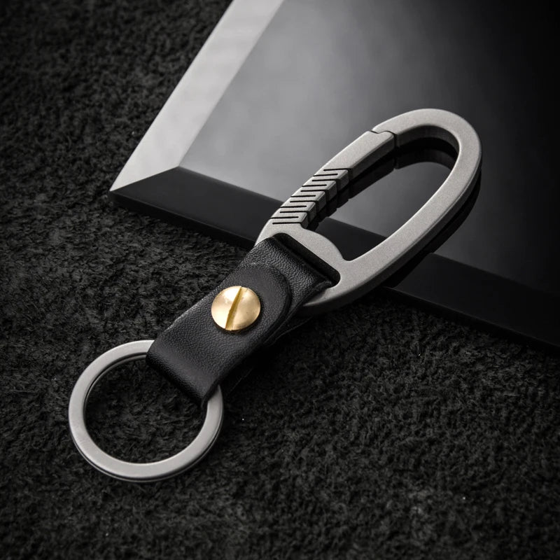 Titanium carabiner with black leather strap and gold accent on a dark textured background.