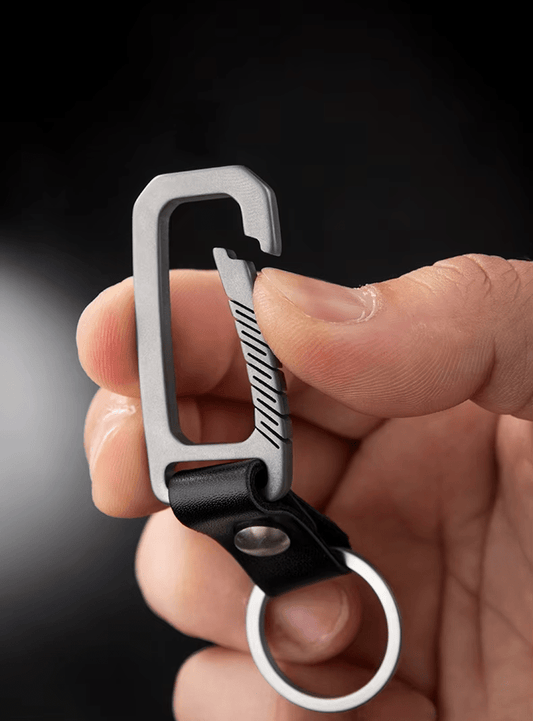 Hand holding a titanium carabiner with a black leather strap against a dark background.